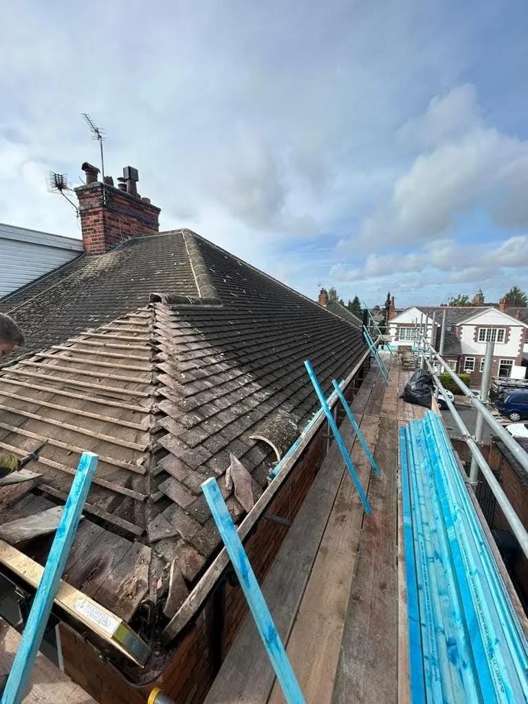 The roof partly stripped: old tiles removed from one hip to expose bare timber battens and boarding underneath, with the rest of the slope still tiled and a two-flue chimney stack behind.