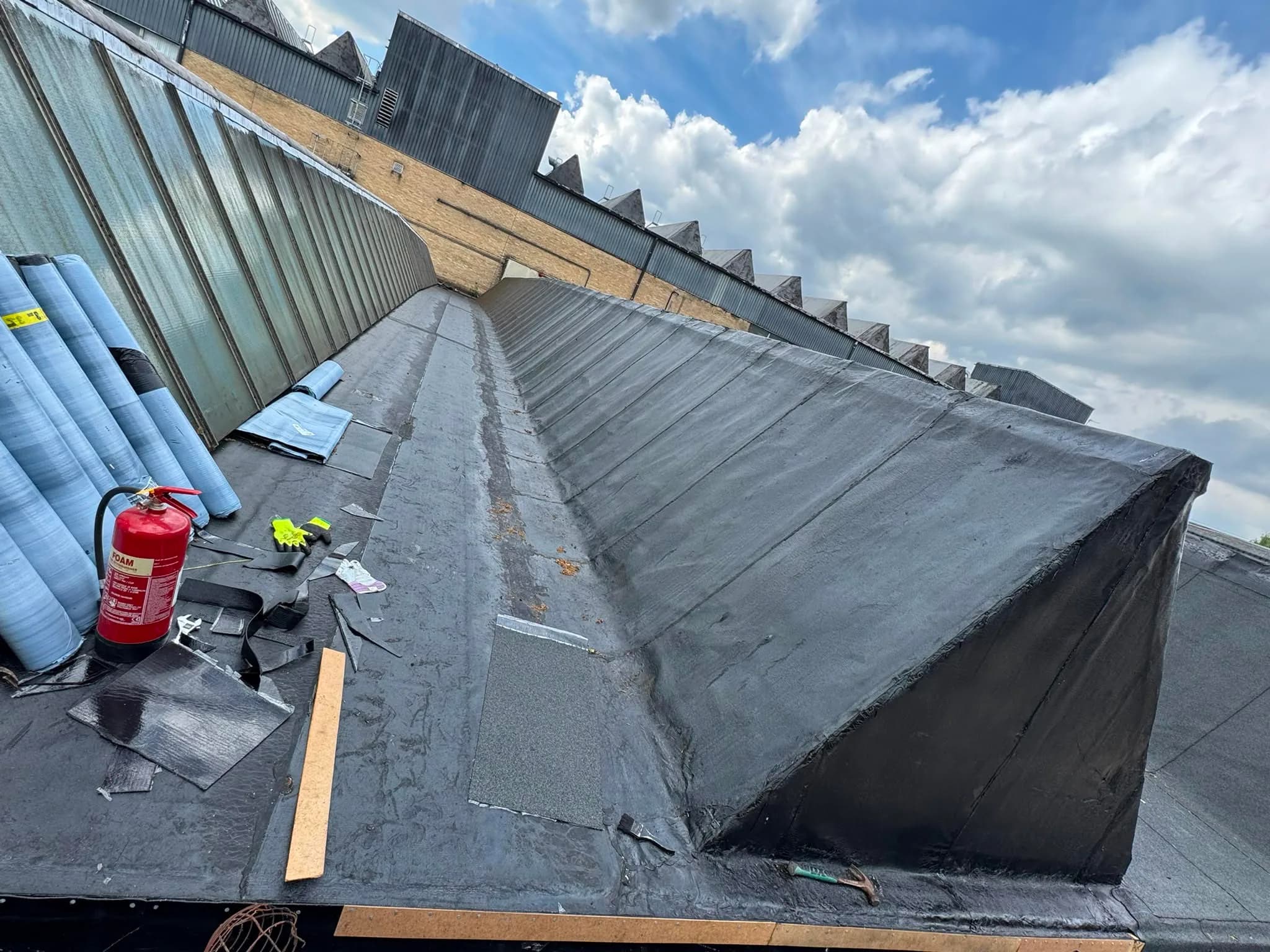 The overlay underway: rolls of underlay stacked on the roof beside a fire extinguisher and hand tools, with the first dark layers of the new bitumen system dressed over a slope and its upstand.