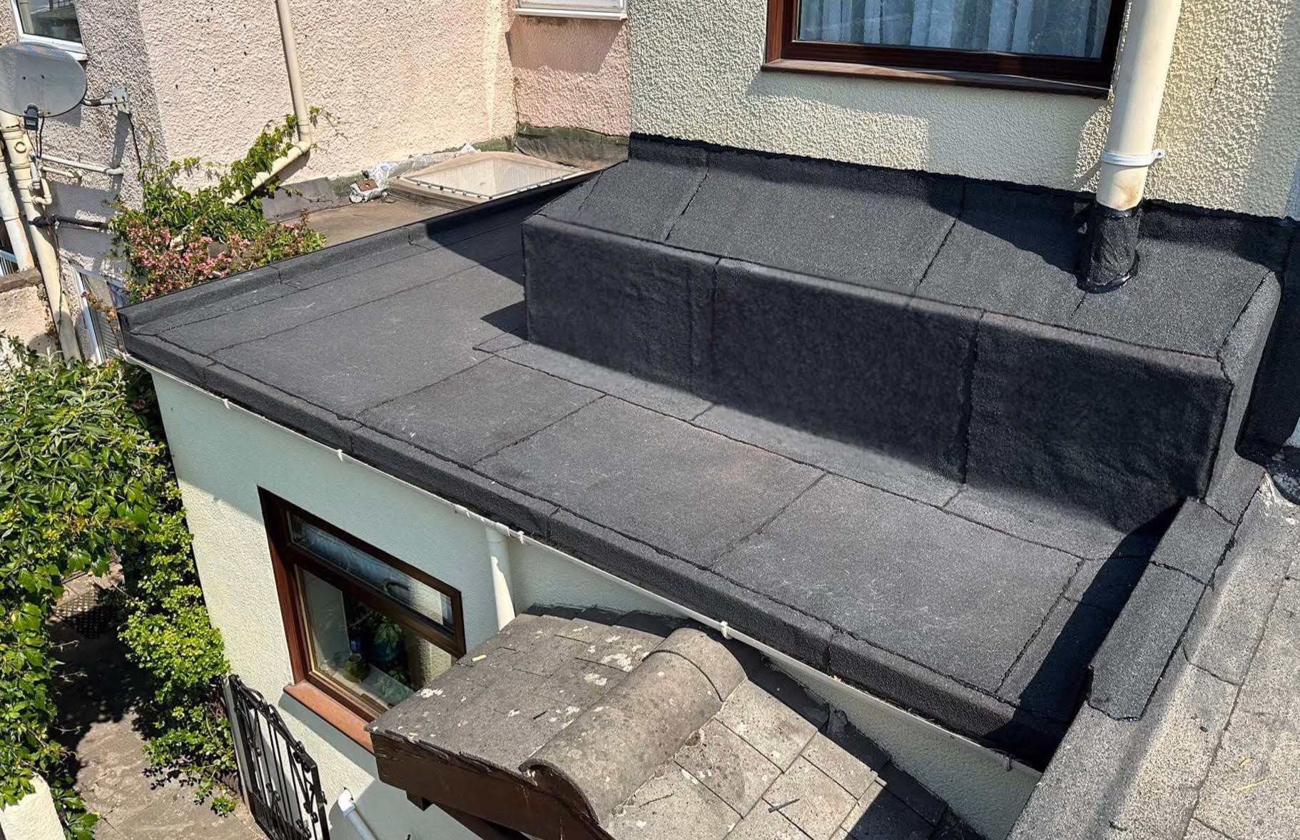 Aerial view of a newly completed flat felt roof with a black mineral-cap finish, showing a raised upstand around a roof window and new lead flashing at a brick chimney stack and soil pipe, against a rendered dormer wall on a terraced house.