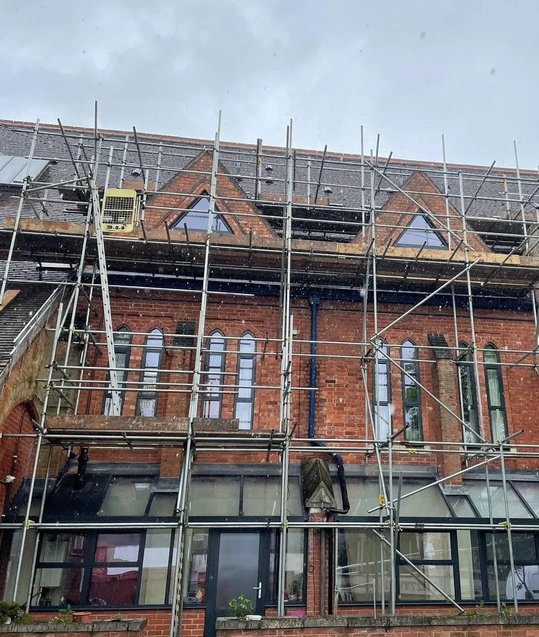A large red-brick heritage building with gothic-arched windows, fully wrapped in scaffolding during roof works in the rain.