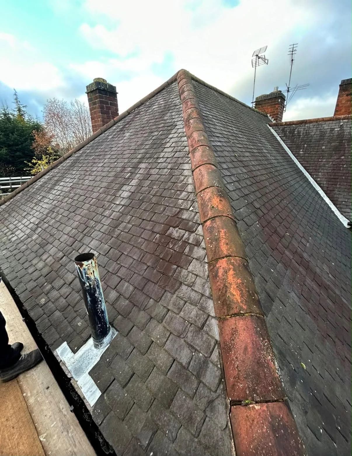 A weathered pitched roof before re-roofing, viewed from roof level: dark, moss-streaked tiles either side of a lichen-covered ridge, with two brick chimney stacks and TV aerials against an overcast sky.