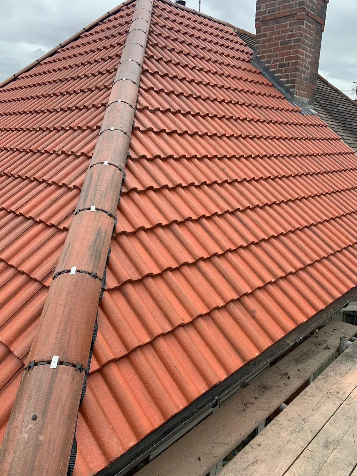 A newly finished orange double-roman tiled roof with a clean ridge line, meeting a brick chimney stack under a grey sky.