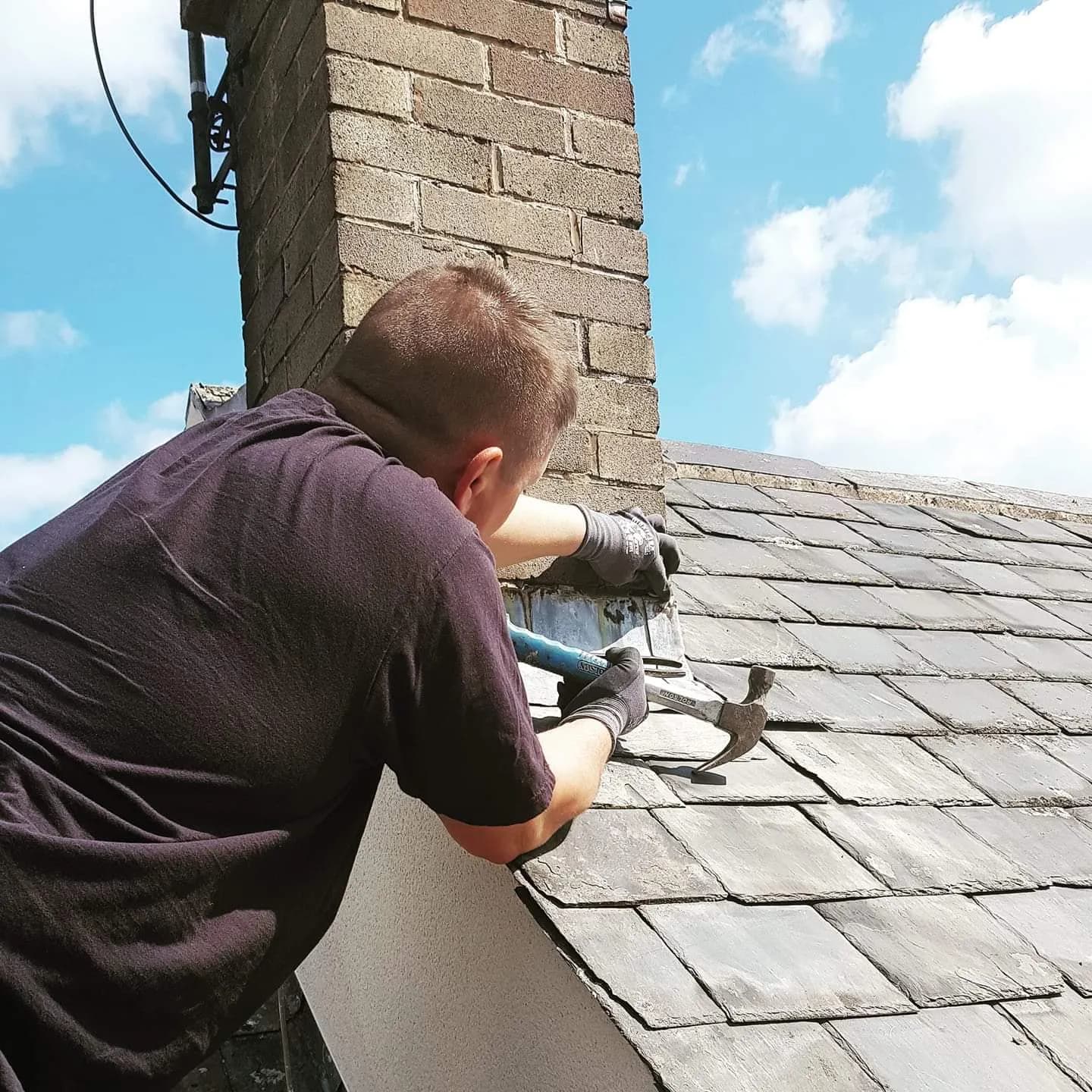 A DLT roofer dressing new lead flashing around the base of a chimney on a natural-slate roof, against a bright blue sky.